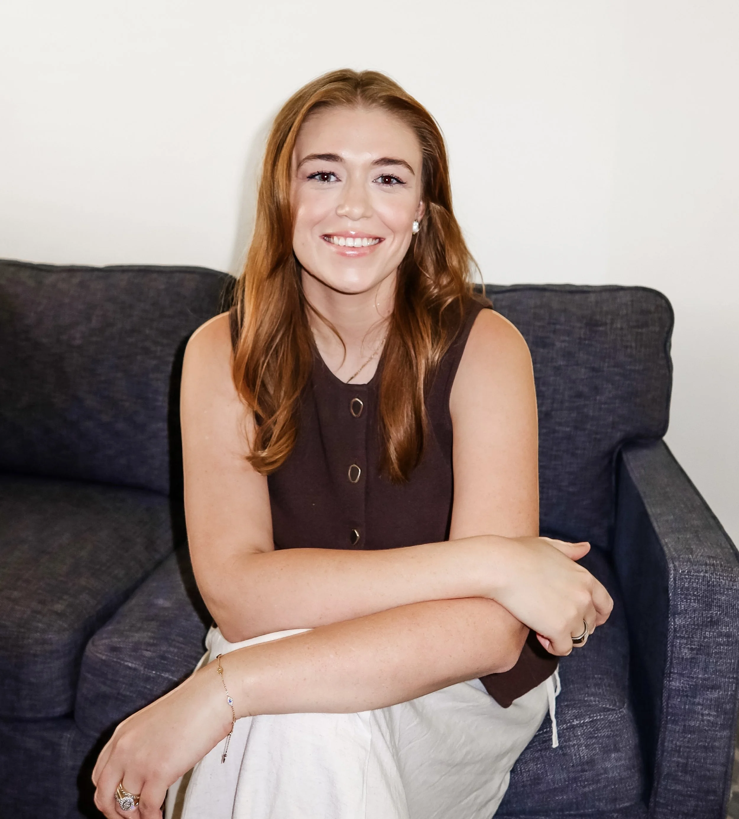 A woman with long red hair sitting on a dark gray couch, smiling at the camera, wearing a sleeveless black top and white pants.