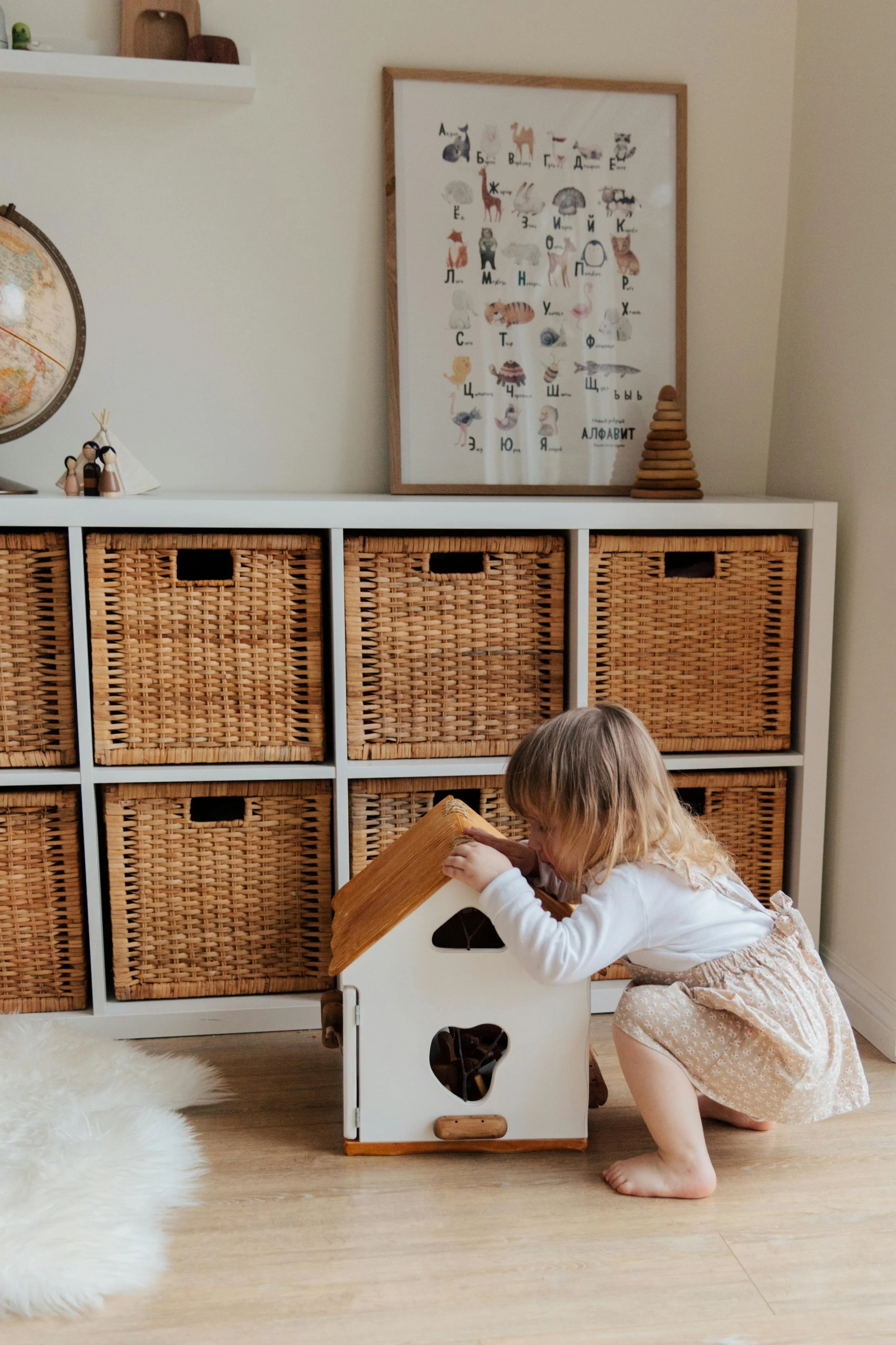 A young girl with blonde hair playing with a wooden dollhouse in a room with wicker storage baskets and a framed animal alphabet poster on the wall.