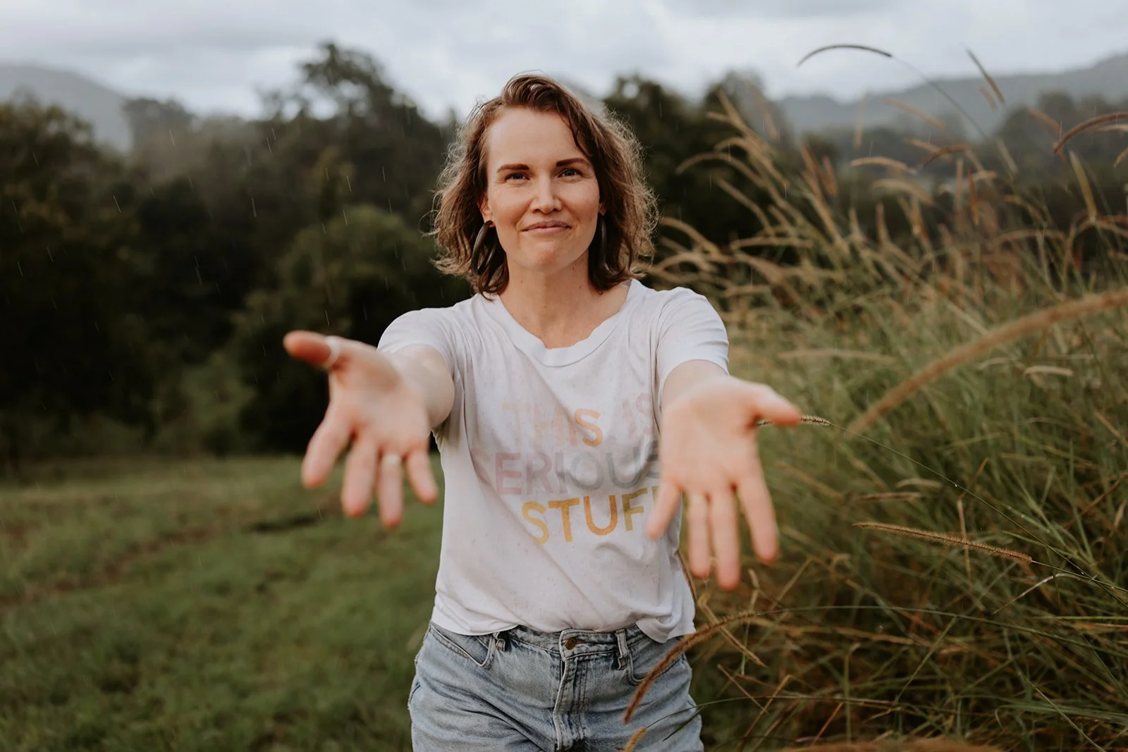 A smiling woman with short brown hair reaching out with both hands outdoors in a grassy field, with tall grass and trees in the background.