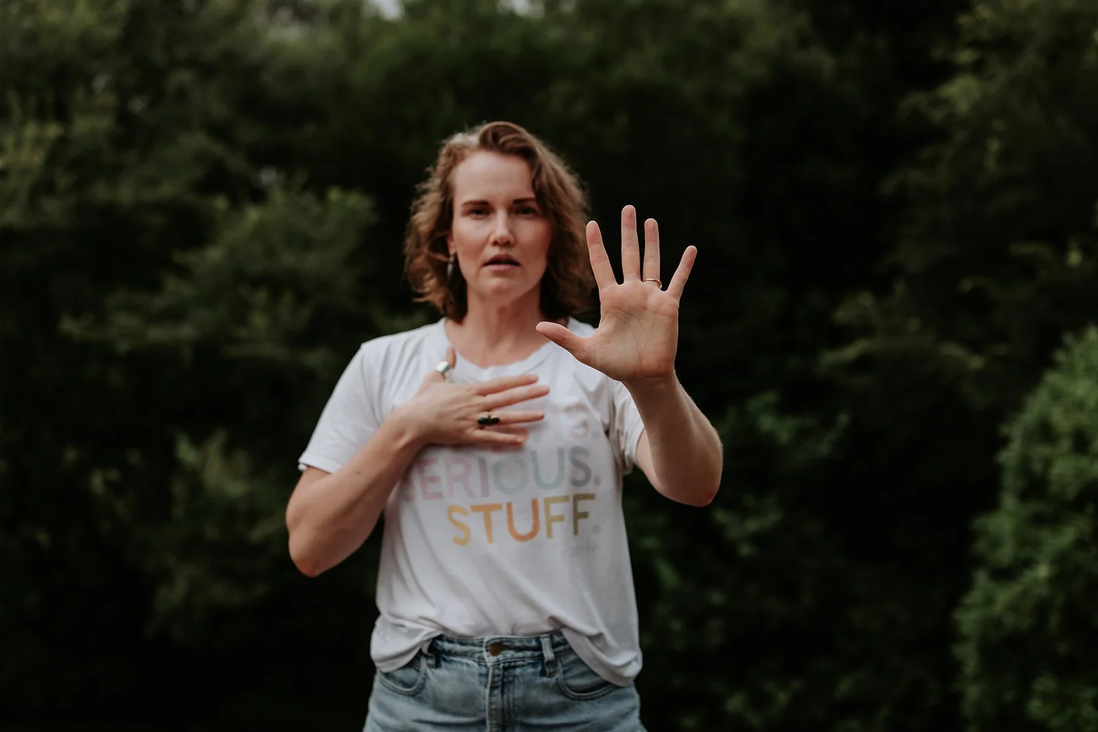 Woman with short, curly hair wearing a white T-shirt with the words 'SERIOUS STUFF' and light denim jeans, standing outdoors with a background of green trees, holding one hand to her chest and raising the other hand in front of her with her palm facing outward.