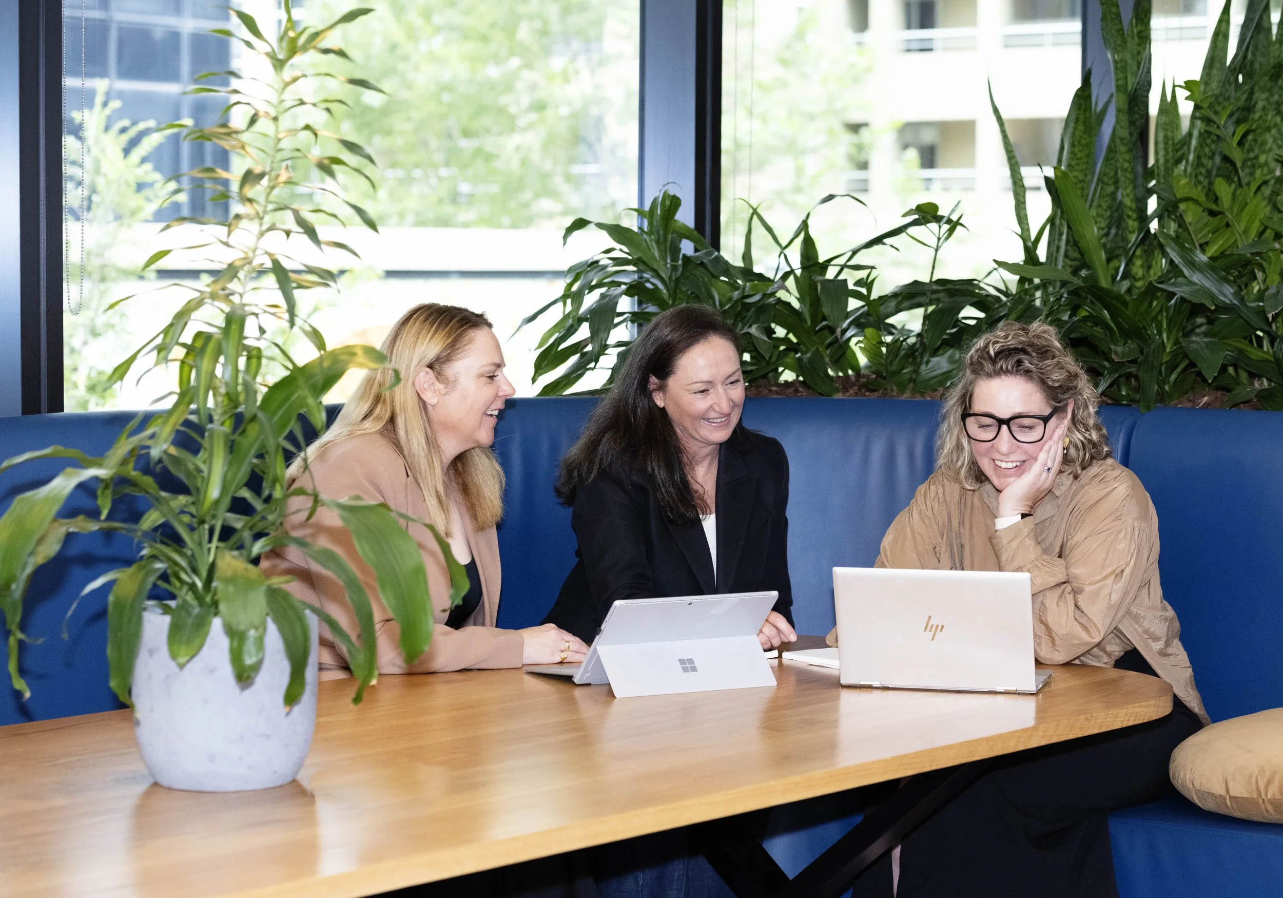 Three women from SP Talent Solutions, sit around a wooden table with laptops and digital tablets, smiling and laughing, in a room with large windows and green plants in the background.