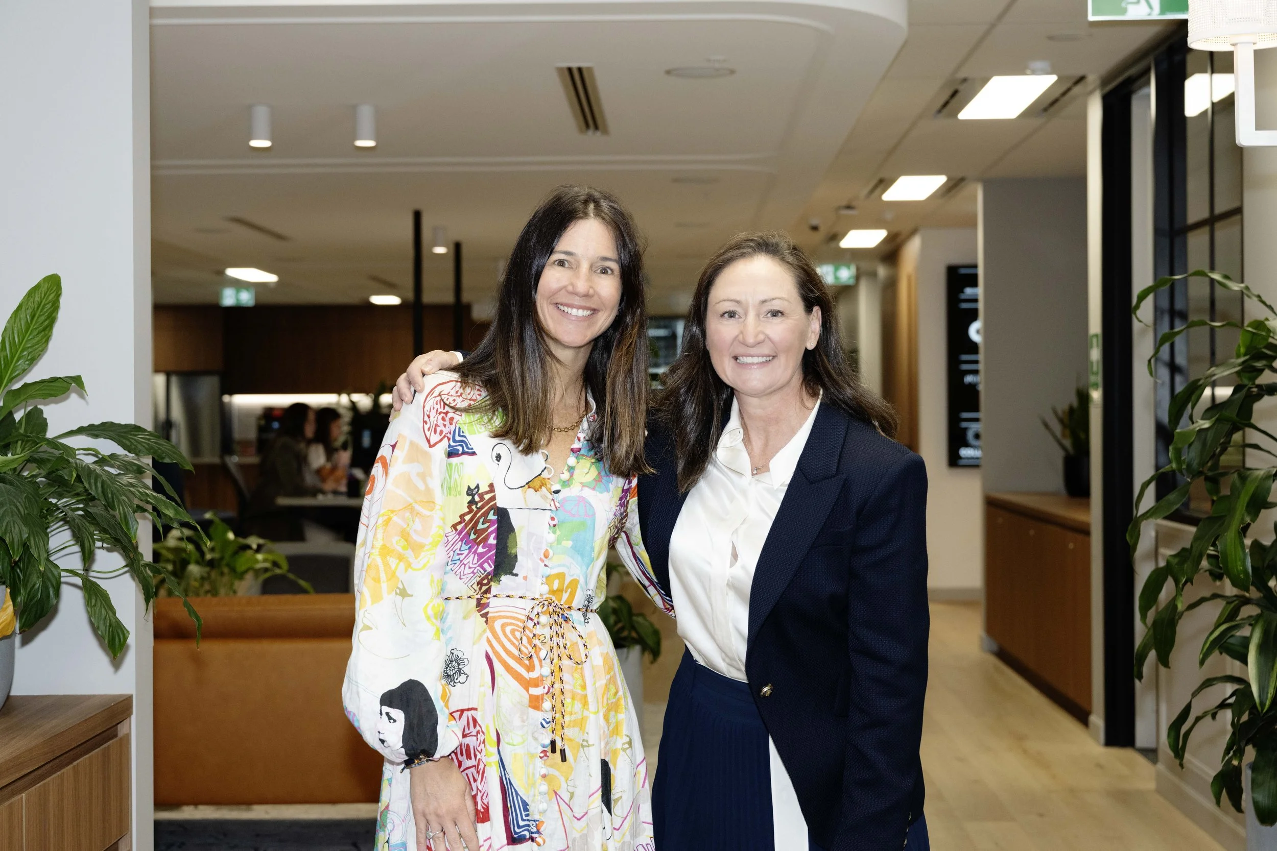 Two women smiling and standing close together inside a modern building, surrounded by green plants, with the woman on the left wearing a colorful patterned dress and the woman on the right wearing a dark blazer and white blouse.