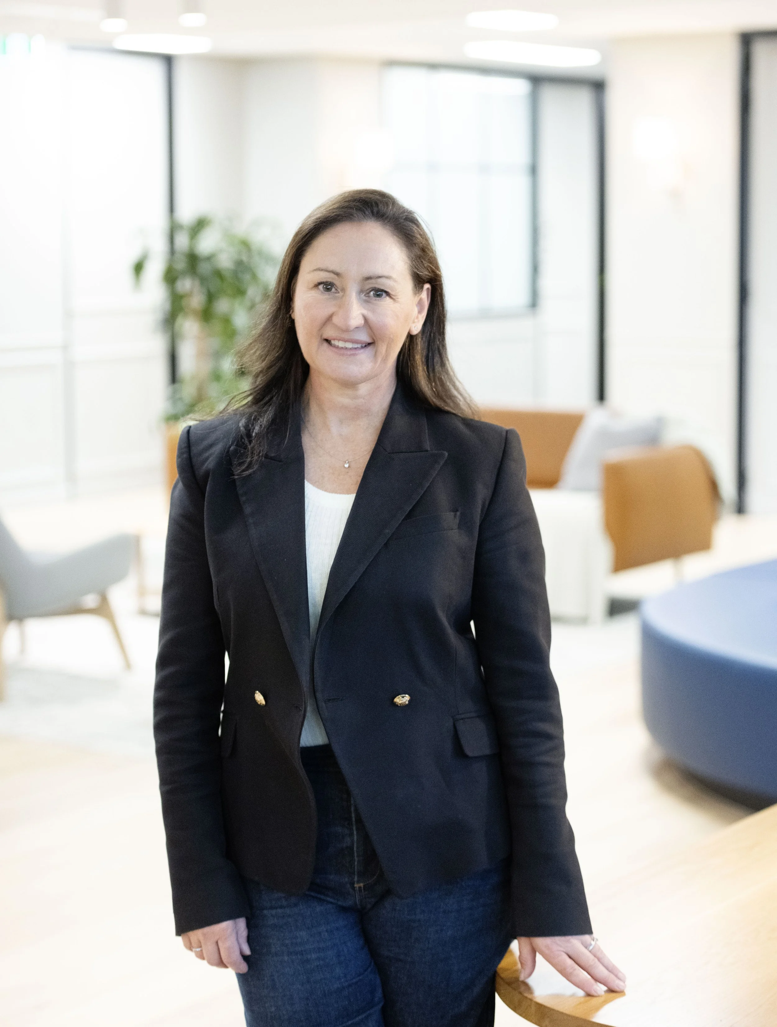 Sarah Pihan of SP Talent Solutions, with long brown hair wearing a black blazer and blue jeans, standing indoors in a modern office or lobby space with glass walls, plants, and chairs. She is smiling and resting her hand on a wooden surface.