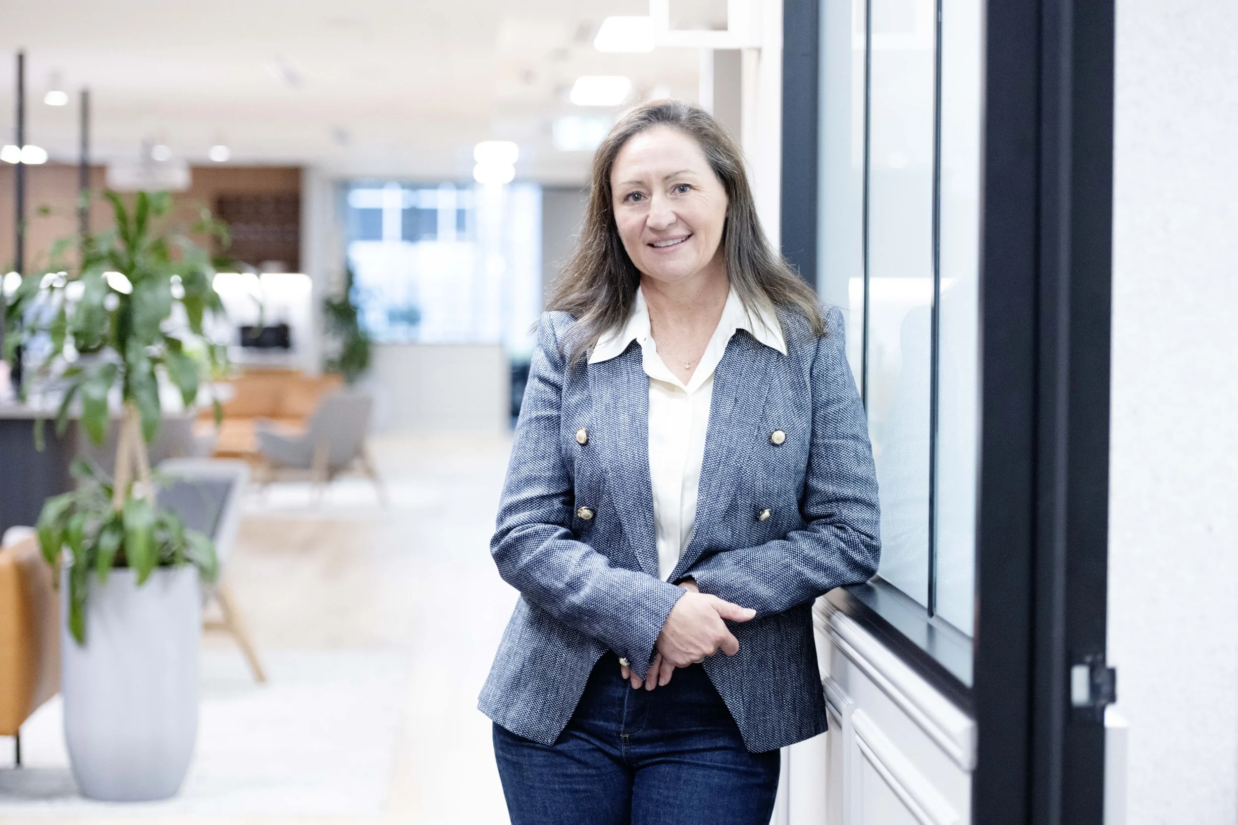 Sarah Pihan of SP Talent Solutions, with long brown hair, wearing a grey blazer over a white shirt, standing indoors near a large window in a modern office or lobby area with plants and seating in the background, smiling and looking to her left.