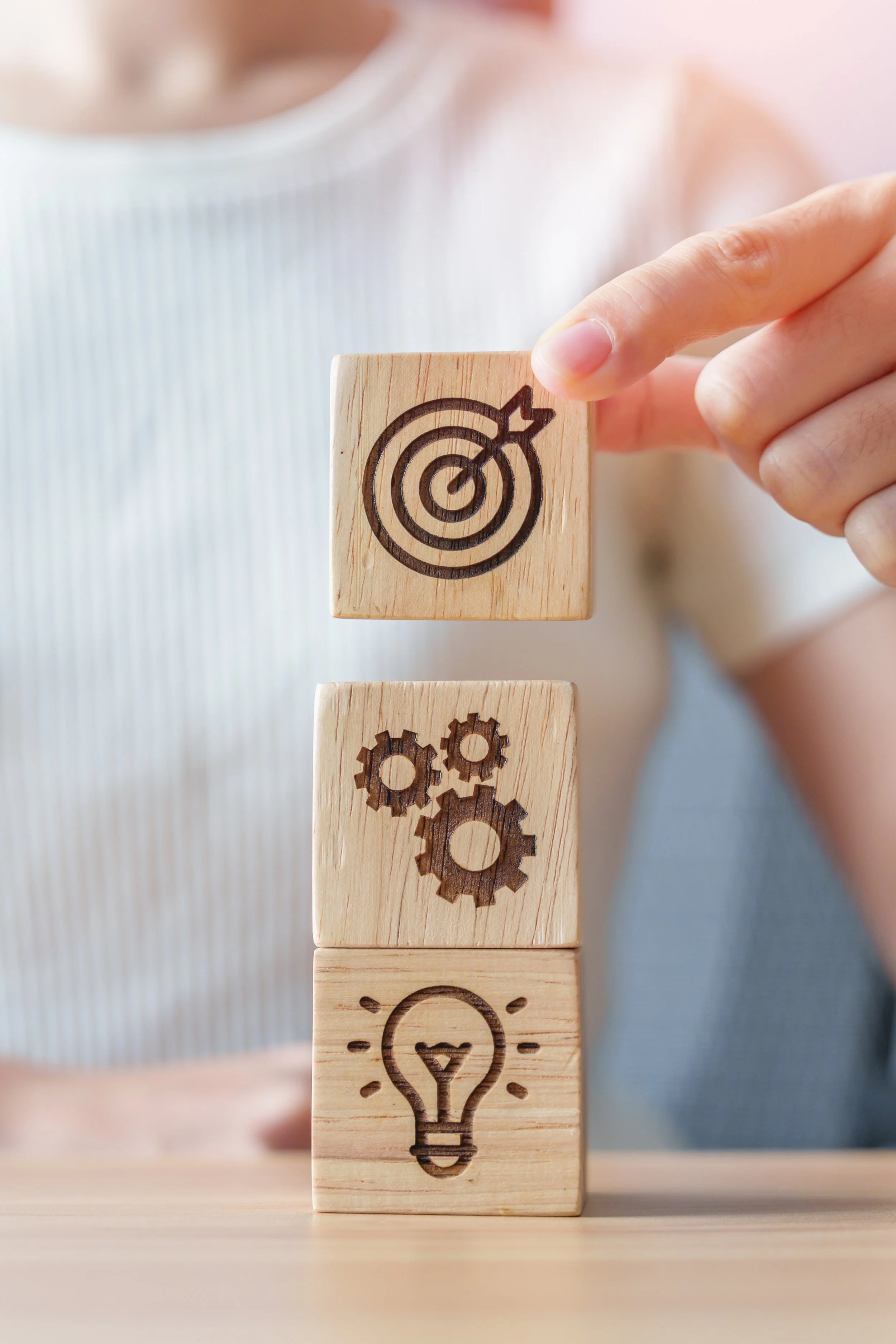 Hand placing three wooden blocks with symbols: a dartboard above a gear above a lightbulb.