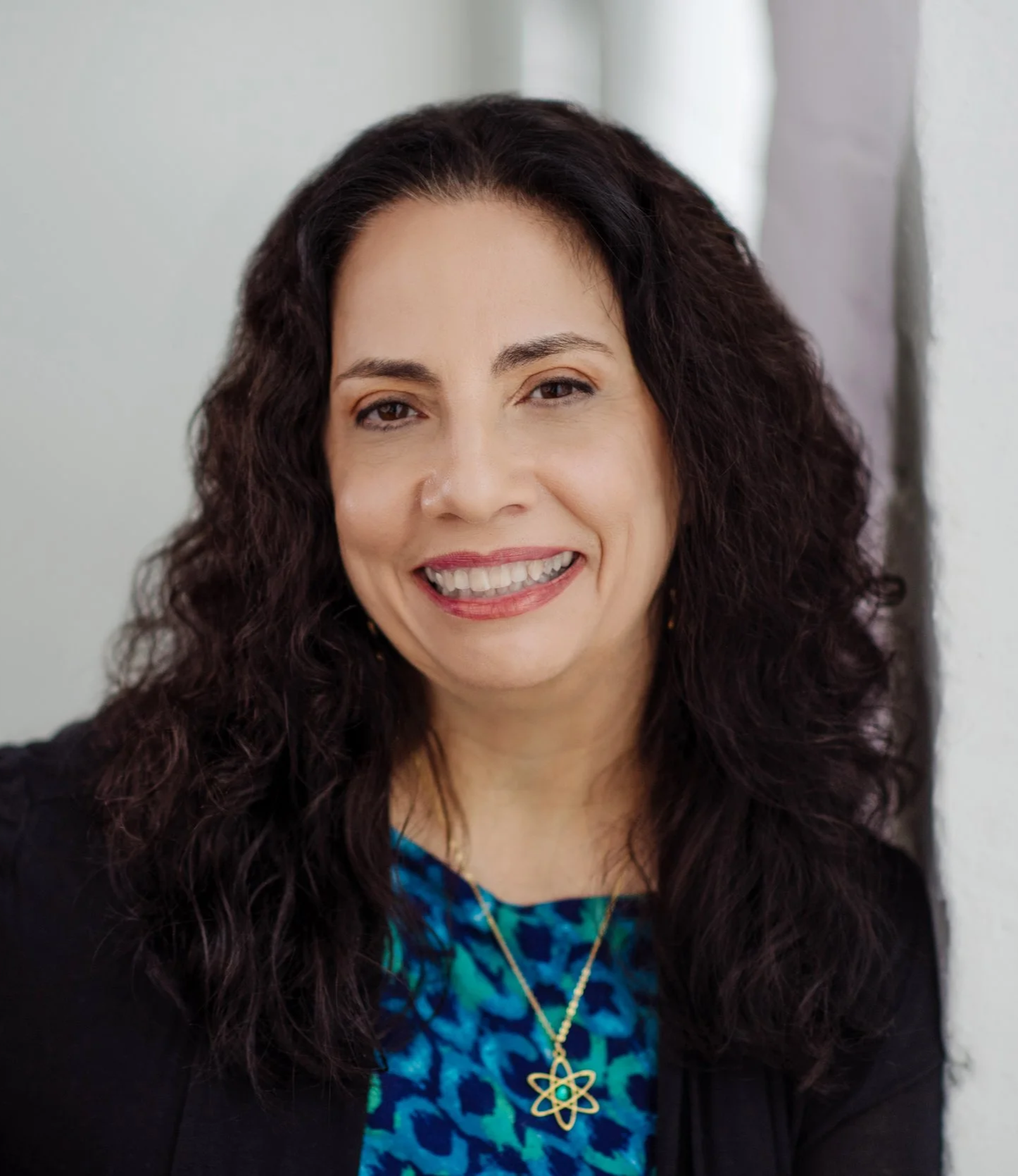 A woman with dark, curly hair smiling at the camera, wearing a blue patterned top and a black blazer, with a background of a plain wall.