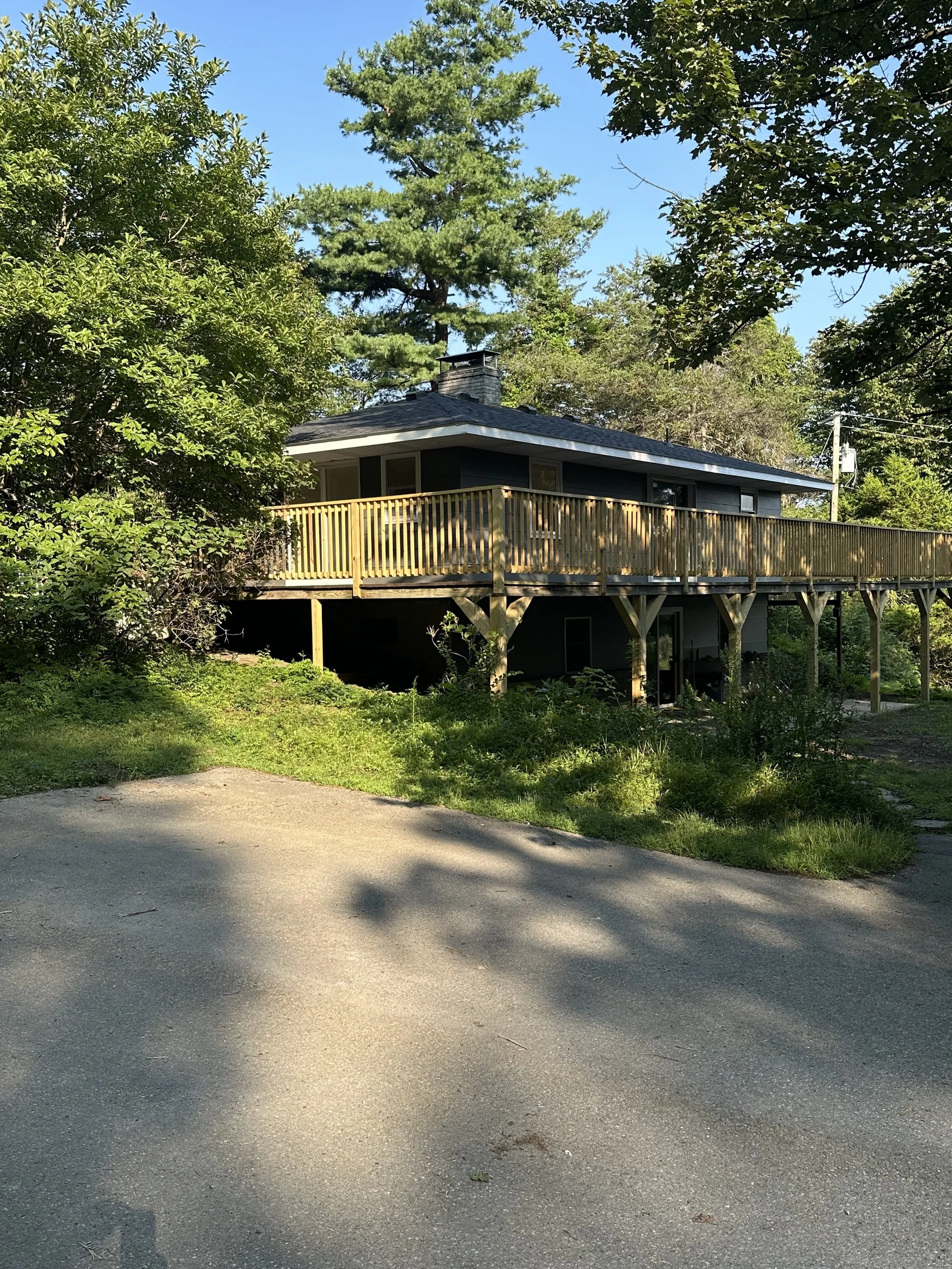 A two-story house with a dark exterior and large wooden deck in a wooded area, surrounded by trees and greenery.
