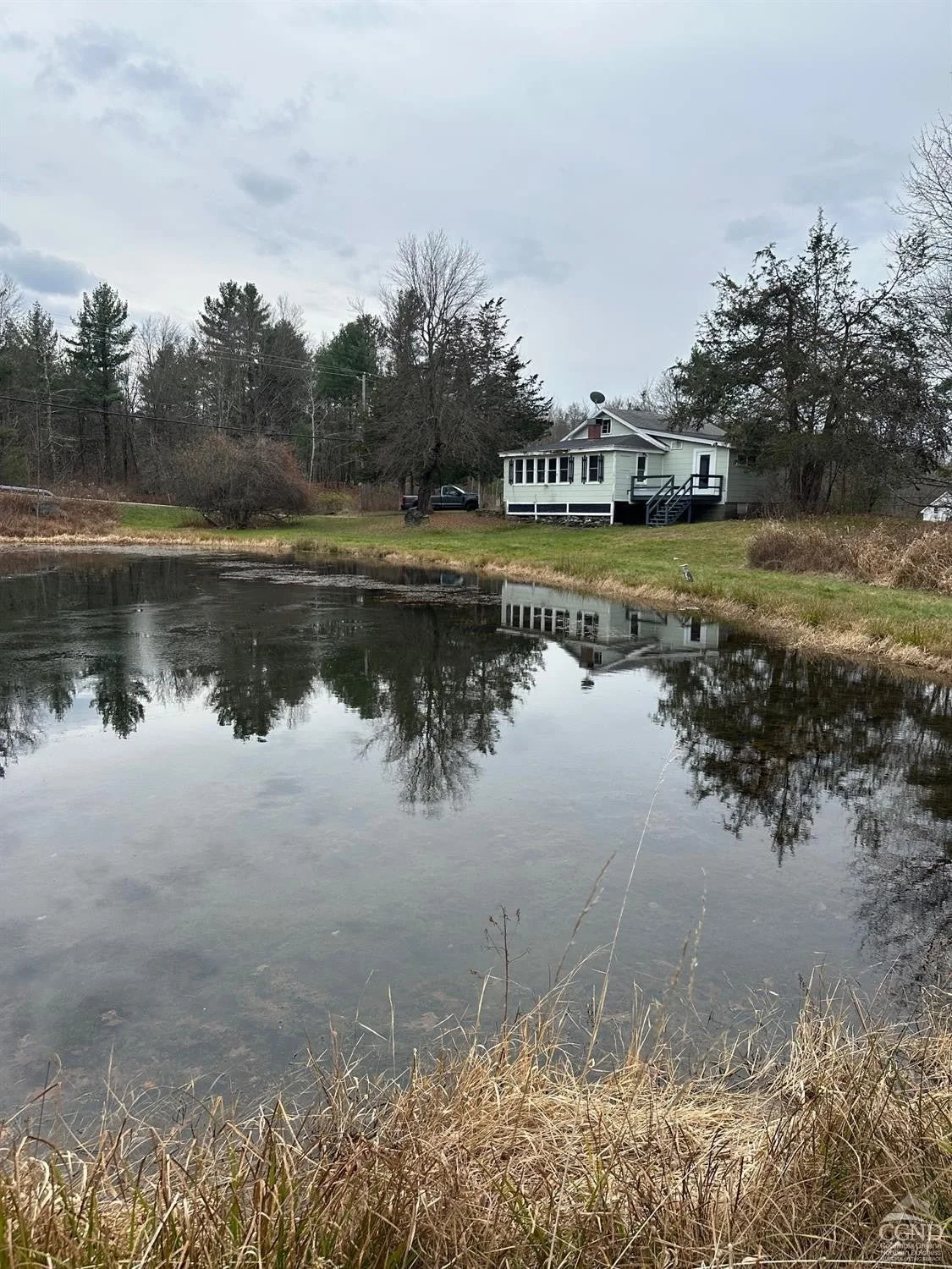 A house with a porch and stairs overlooking a pond with trees reflected in the water under a cloudy sky.
