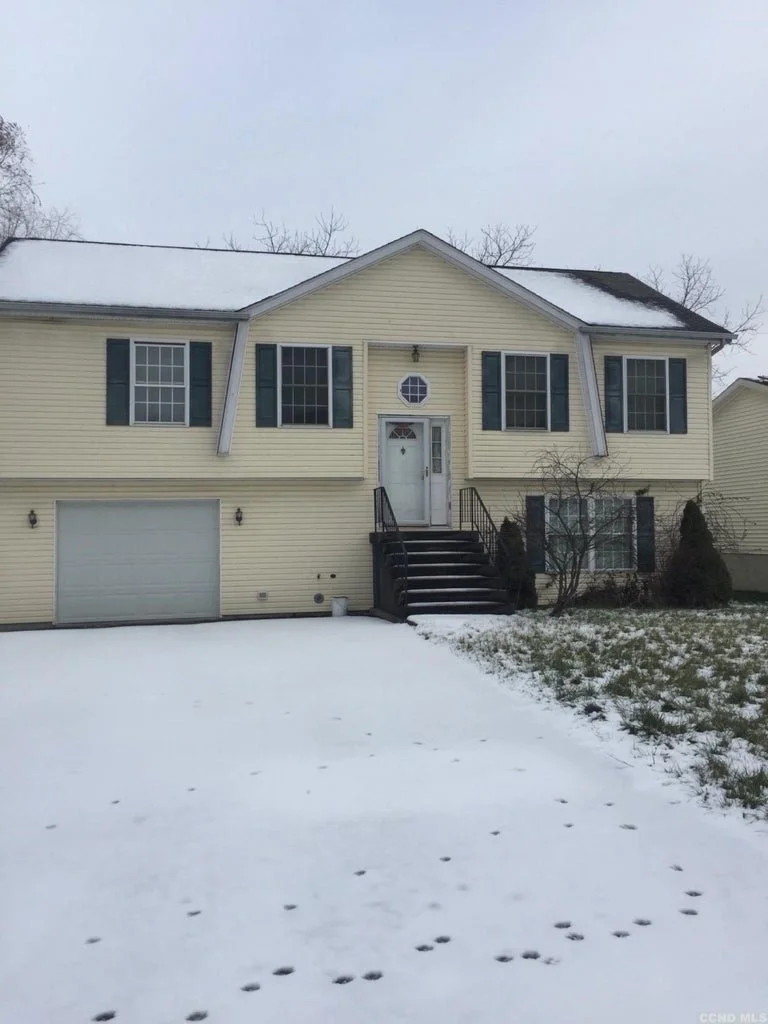 A yellow two-story house with black shutters, stairs leading to the front door, and a garage in a snowy yard.