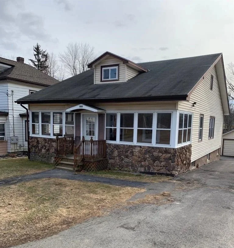 A two-story house with a stone foundation, beige siding, a black roof, and a small porch with wooden stairs. It has large front windows and a small dormer window on the roof.