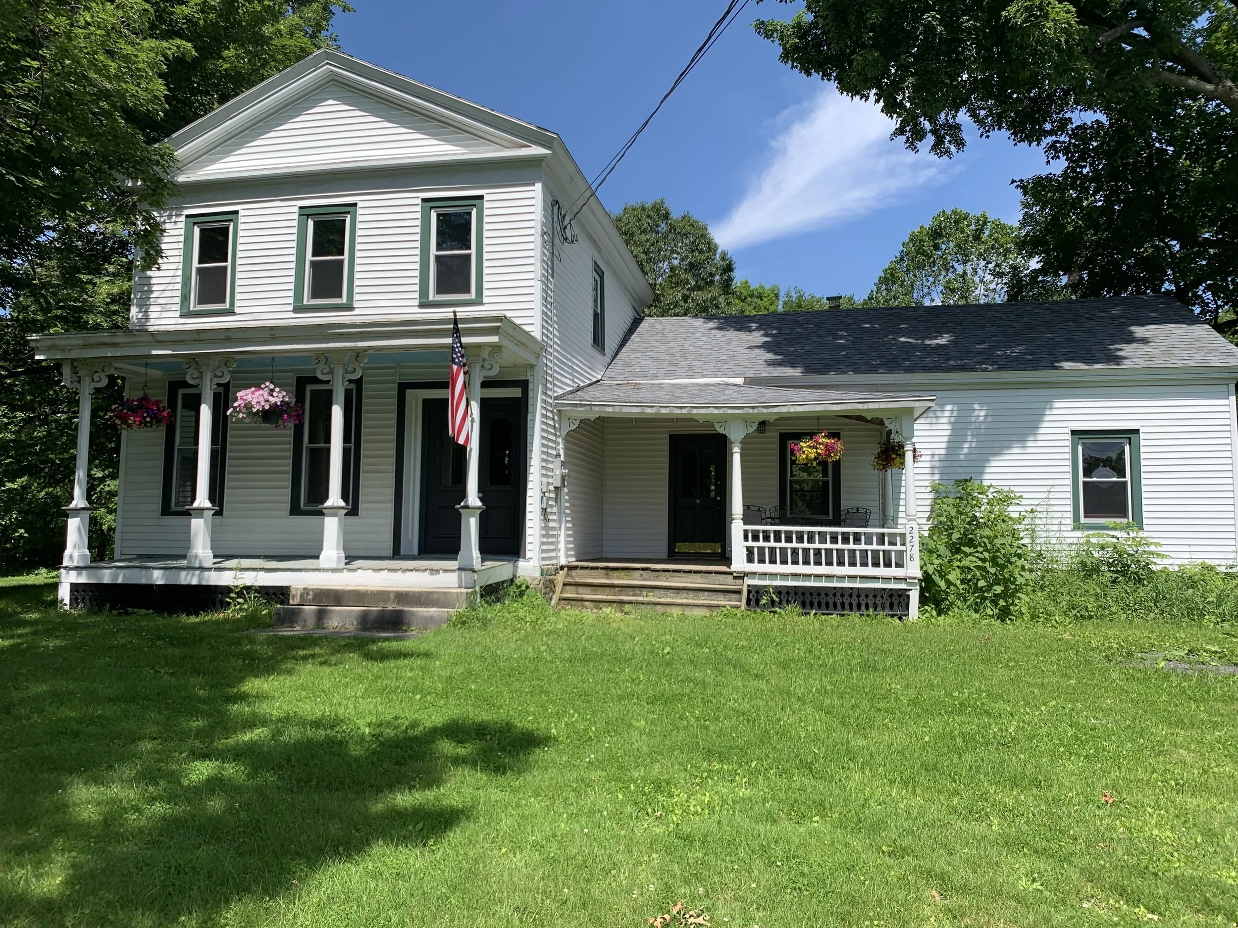 White old-fashioned house with front porch, hanging flower baskets, American flag, and green lawn, with trees and blue sky in the background.