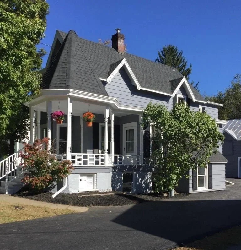 A two-story house painted in light gray with white trim, featuring a front porch with white railings and columns, hanging flower baskets, a dark gray shingled roof, and surrounded by trees under a clear blue sky.