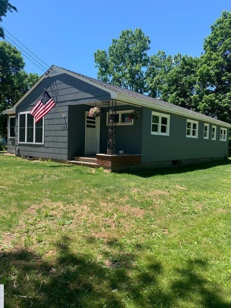 Gray single-story house with a front porch and steps, American flag, hanging flower baskets, and multiple windows under a clear blue sky.
