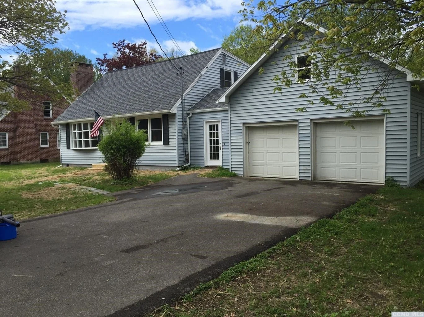 A light blue house with white garage doors and a small front porch, a bush in front, and an American flag near the windows. There is a driveway with some patches and a tree with green leaves overhead.