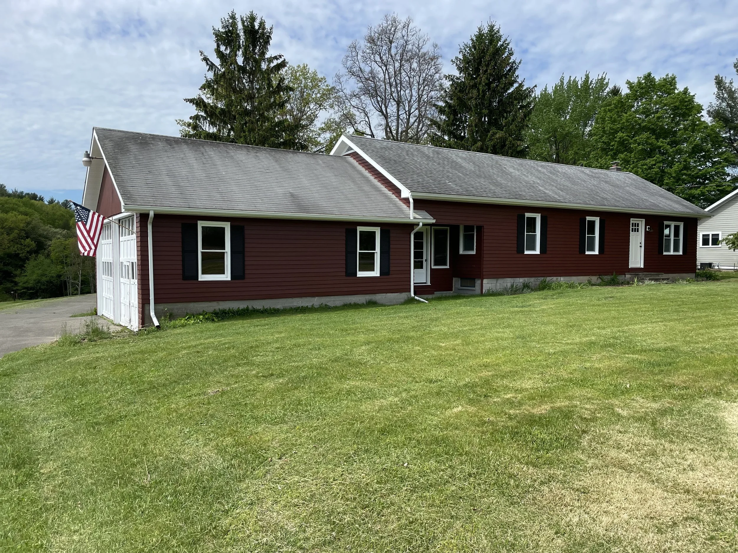 A single-story house painted red with black shutters and white trim, with a attached garage on the left side and a well-maintained green lawn in front. The house has a gray shingled roof, and the sky above is partly cloudy. An American flag is mounted near the garage.
