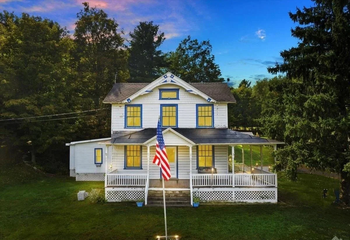 A large white two-story house with blue trim and yellow windows, front porch, American flag on a pole, surrounded by trees at dusk.