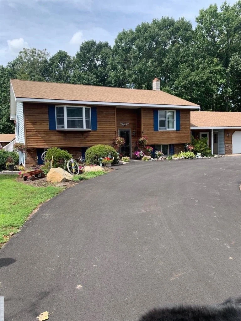 A two-story house with wood siding, blue shutters, a white roof, a small front porch with flowers, and a driveway with plants and decorations in the yard, surrounded by trees.