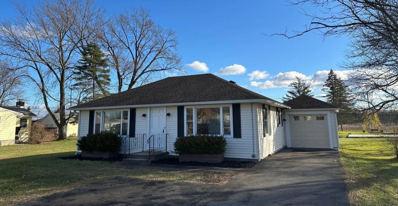 A single-story house with white siding, a dark shingled roof, and blue shutters. There are bushes in front and a driveway leading to a garage on the right. The yard is grassy, and trees are visible in the background with a partly cloudy sky.