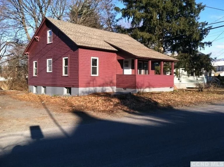Red house with a porch and multiple windows, situated on a partially grassy lot with trees in the background, casting shadows on the street during daytime.
