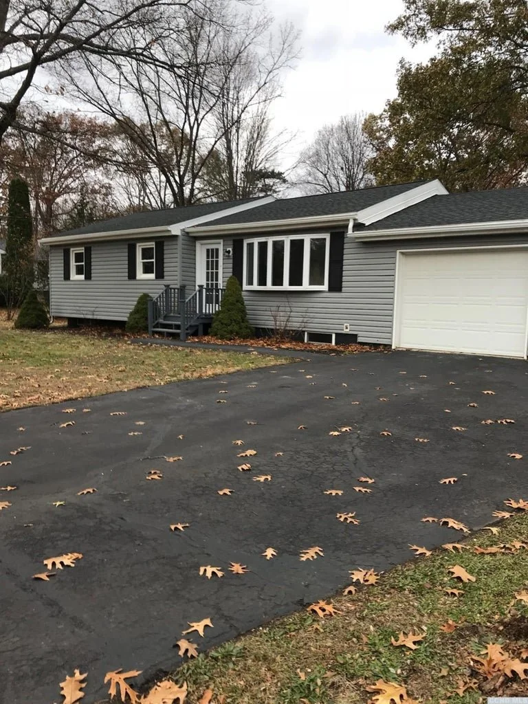 A gray house with black shutters and a white garage door, surrounded by a paved driveway with scattered fallen leaves, and trees with bare branches in the background.
