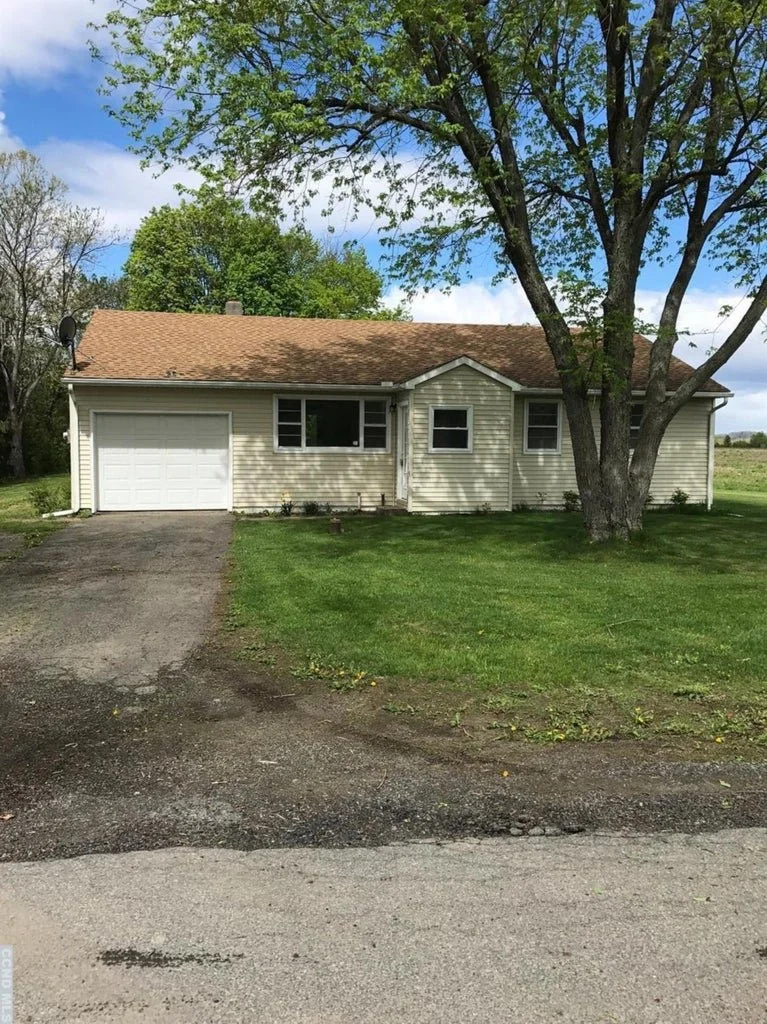 A single-story house with beige siding, a brown shingled roof, and a white garage door. There is a large tree in the front yard with green leaves, and the yard has green grass. The driveway is made of asphalt, and the sky is partly cloudy with some blue showing.