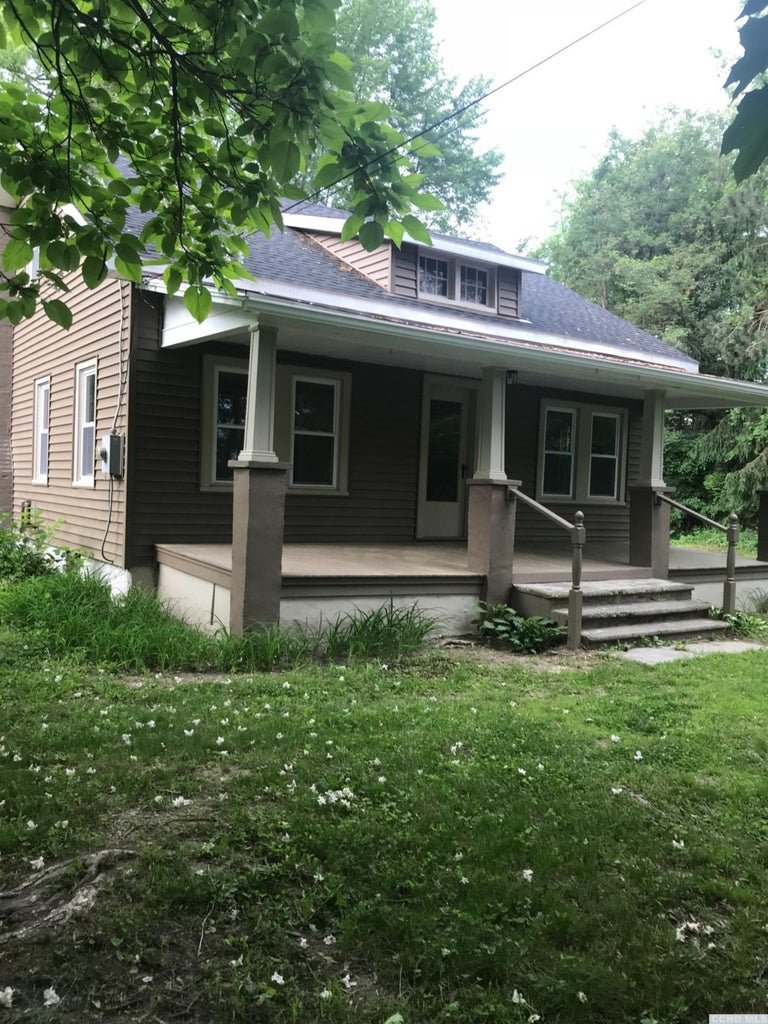 A house with brown siding, white trim, and a front porch with steps, surrounded by grass and trees.