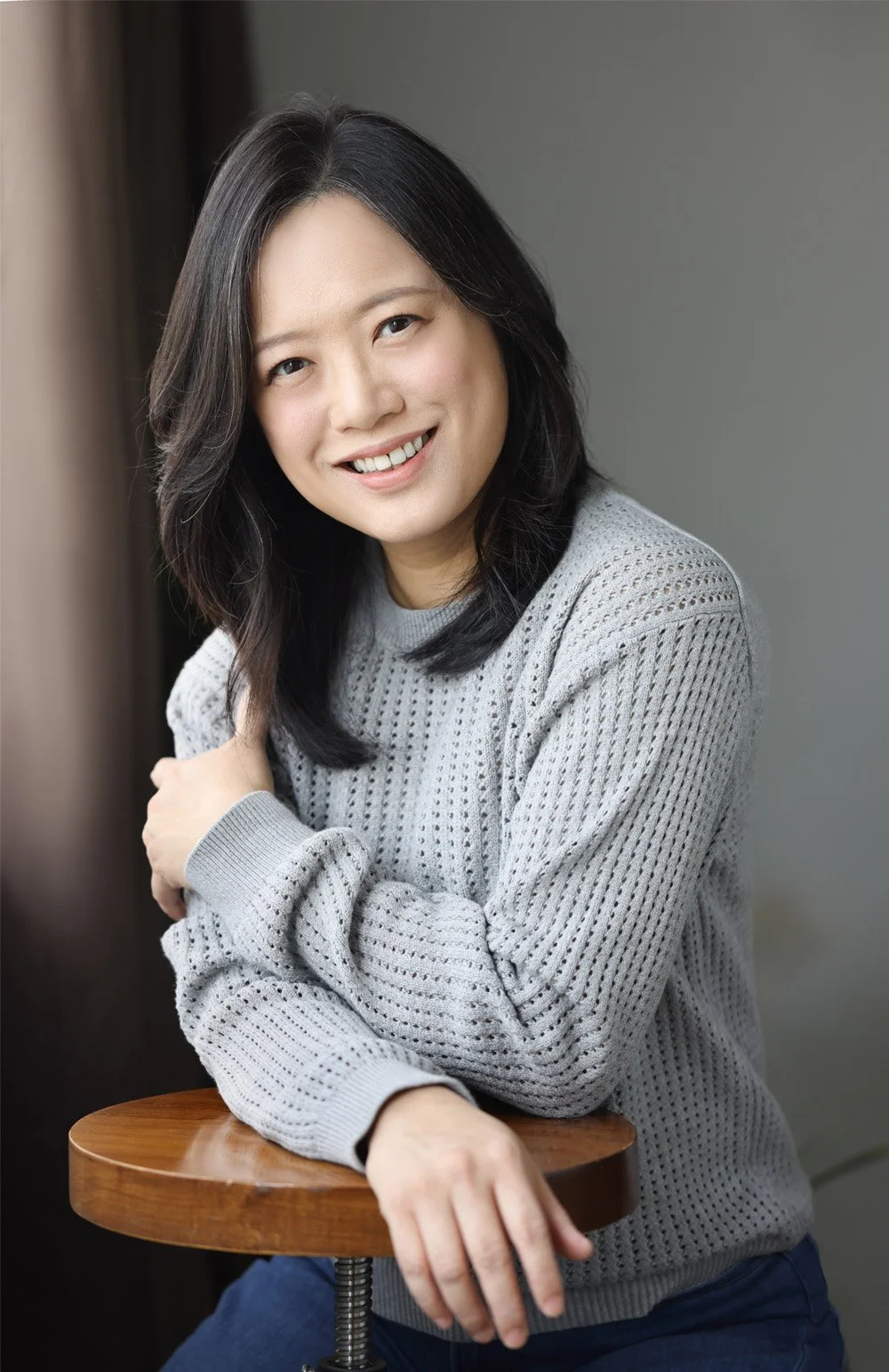 A woman with shoulder-length black hair, wearing a light gray cable-knit sweater, sitting at a wooden table with her arms crossed on top of the table, smiling and leaning slightly forward in a relaxed pose.