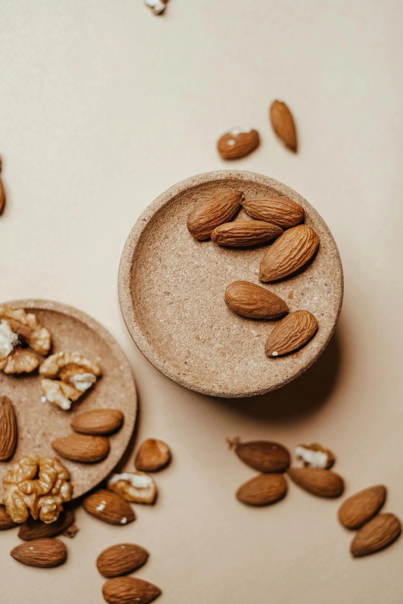 Almonds and walnuts in small, round, beige dishes on a neutral background.