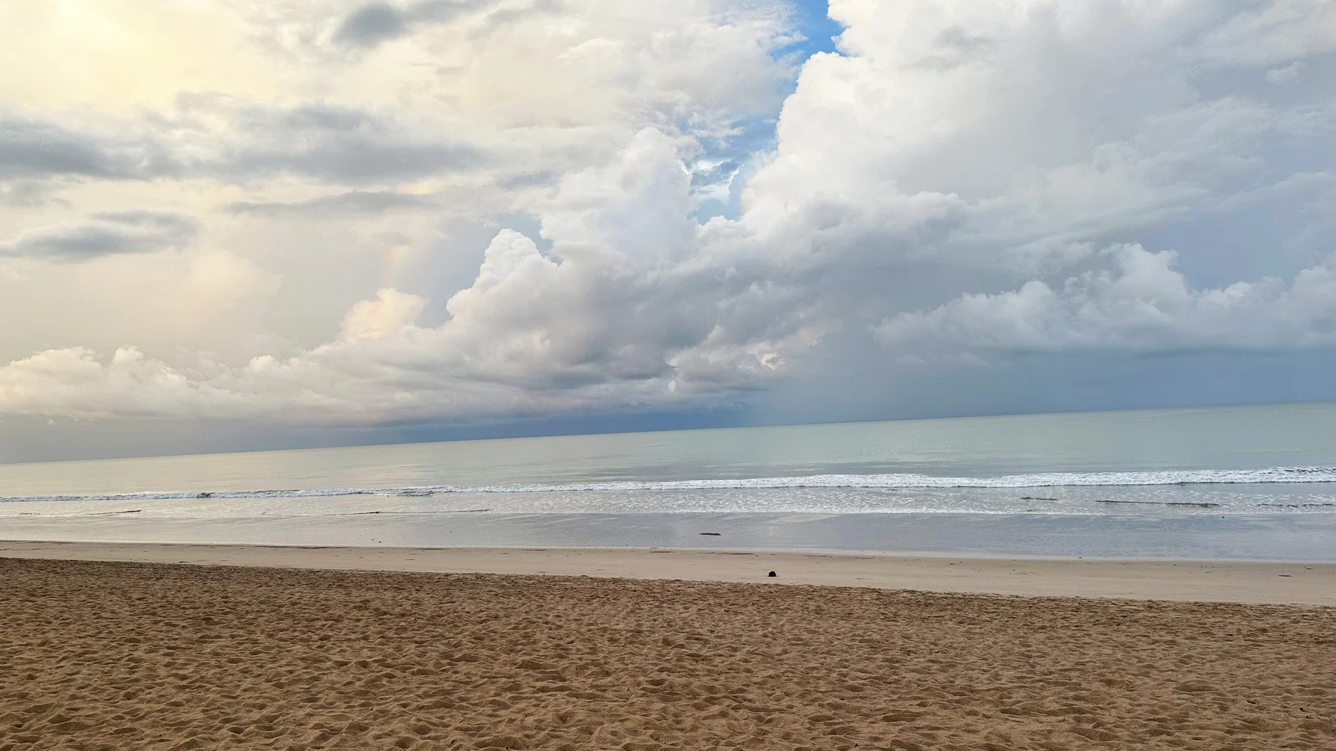 A sandy beach with calm ocean waves under a cloudy sky.