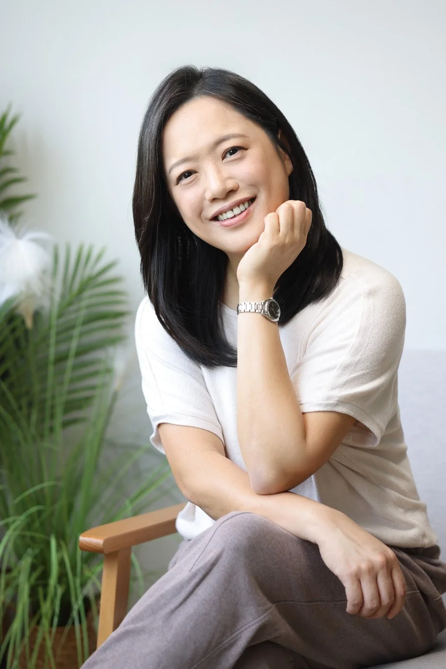 A woman with shoulder-length dark hair smiling, sitting indoors with green plants in the background, wearing a white t-shirt and a silver watch.
