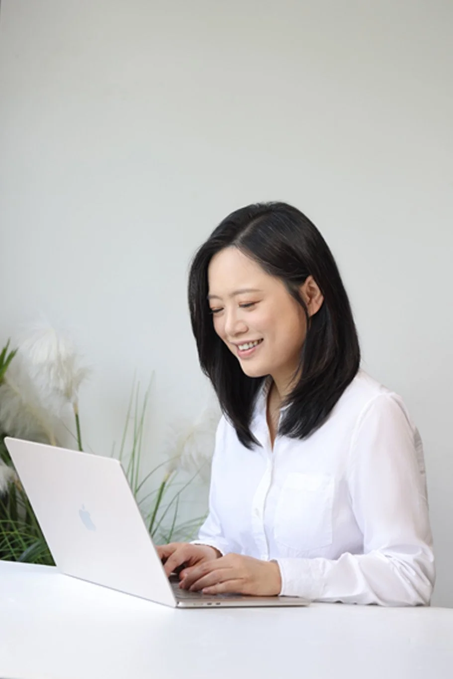 Young woman with black hair smiling and using a silver MacBook laptop at a white desk, with white flowers and green plants in the background.