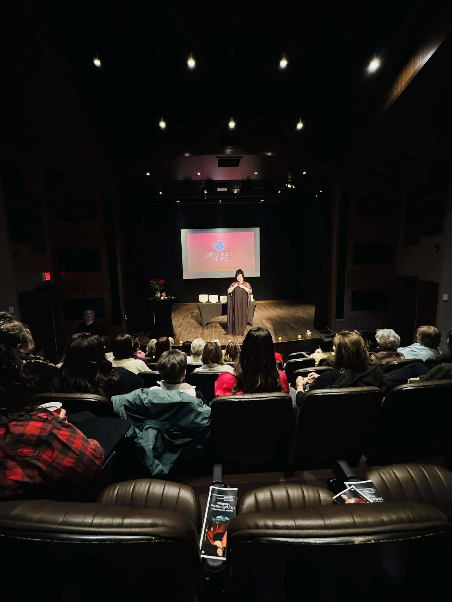 An audience watching a woman speak on stage in a theater. The stage has a screen displaying a presentation with the name 'Michelle Munk'.