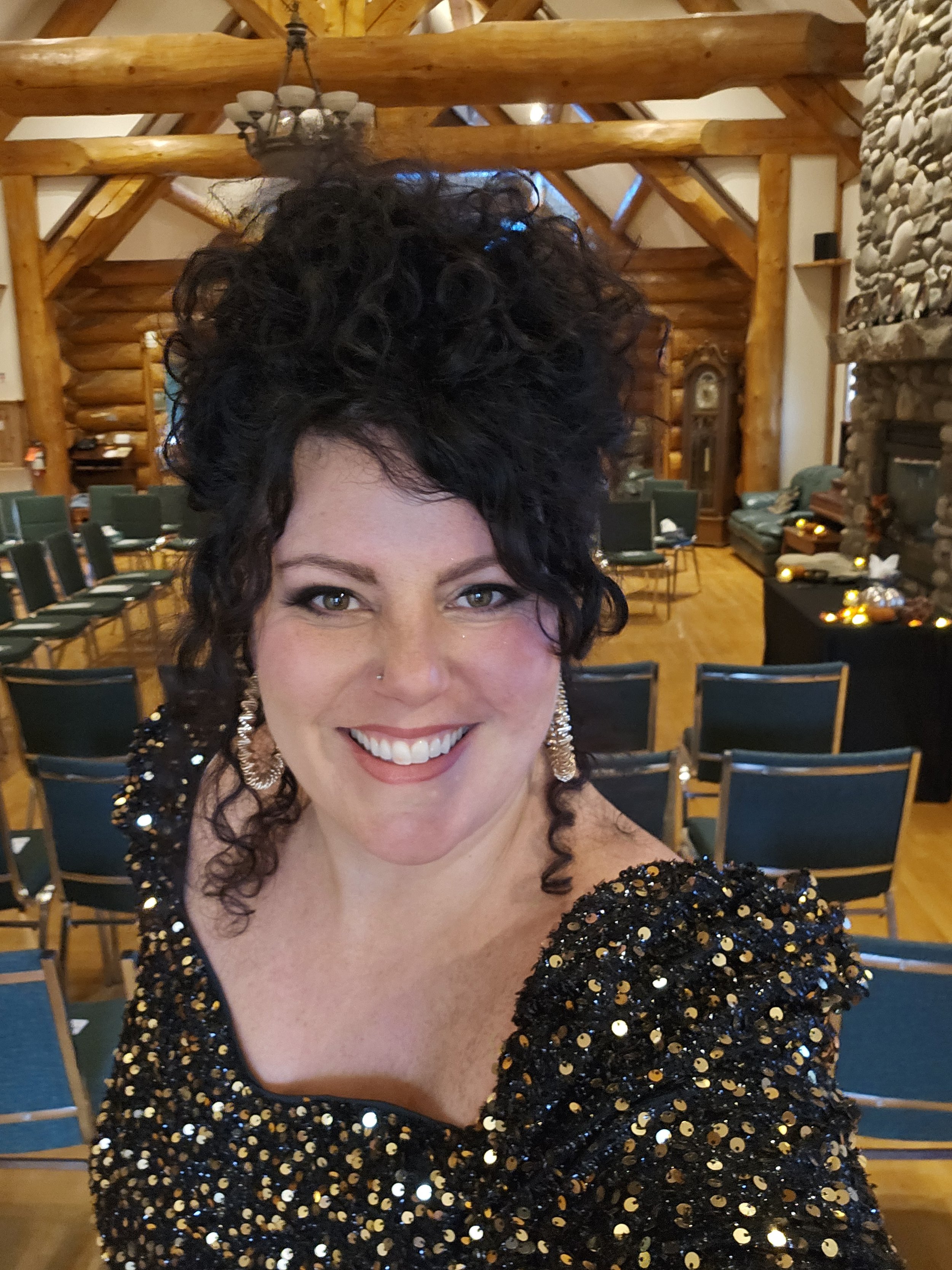 Smiling woman with dark curly hair, wearing a black sequin dress and earrings, in a rustic log cabin event room.