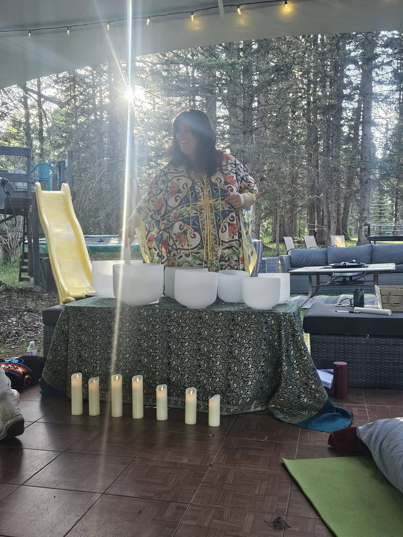 A woman standing at a table with white singing bowls outdoors with trees and string lights in the background. Candles are arranged on the floor in front of the table.