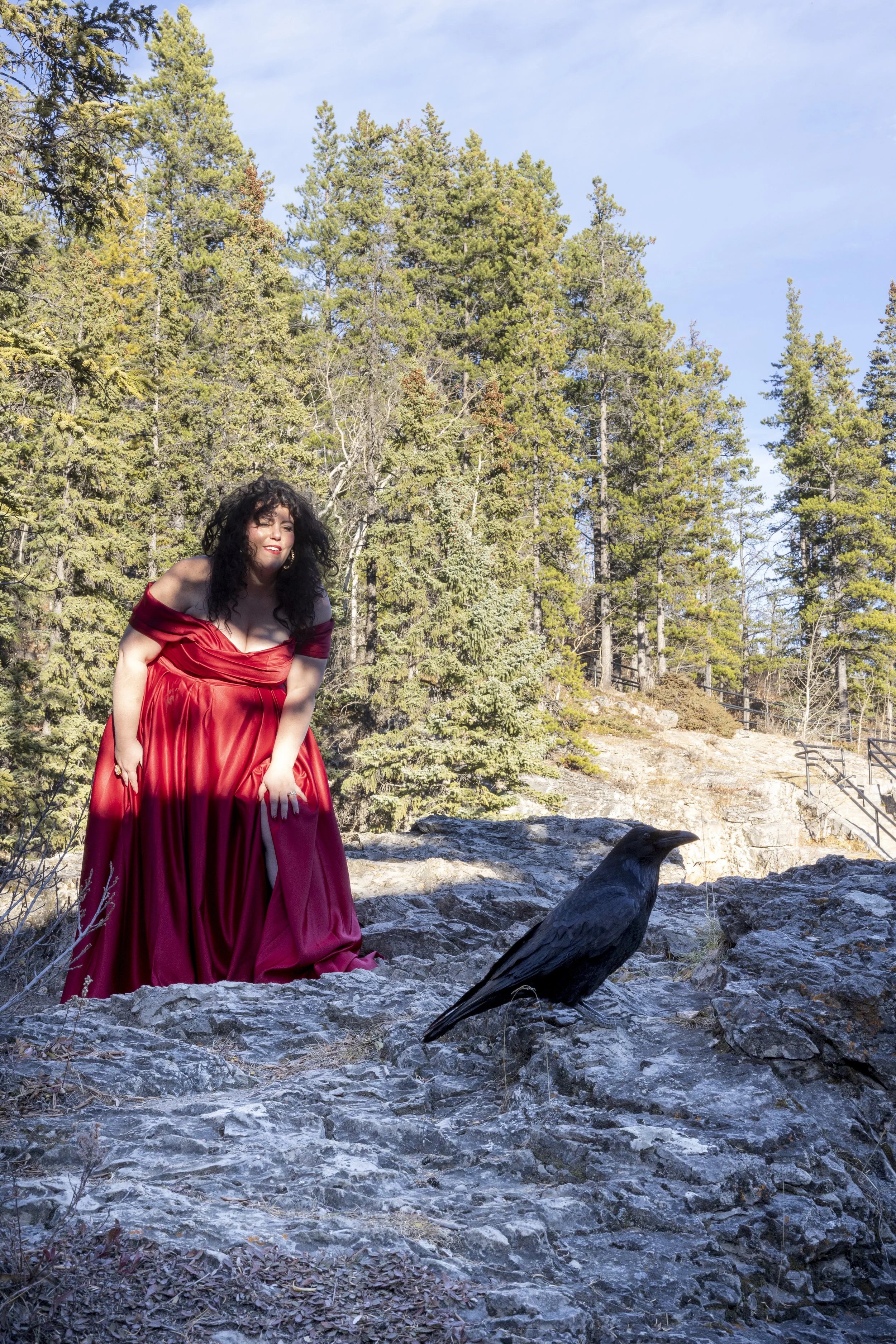 A woman with long dark curly hair wearing a flowing, off-shoulder red dress standing on rocky ground near a black raven in a wooded outdoor area with tall trees and a clear blue sky.