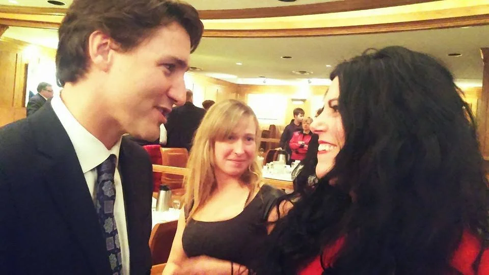 Three people inside a restaurant or banquet hall. A young man in a suit and tie facing a woman with long black hair and a red top. A woman with blonde hair and black top is in the background, smiling and looking at them.