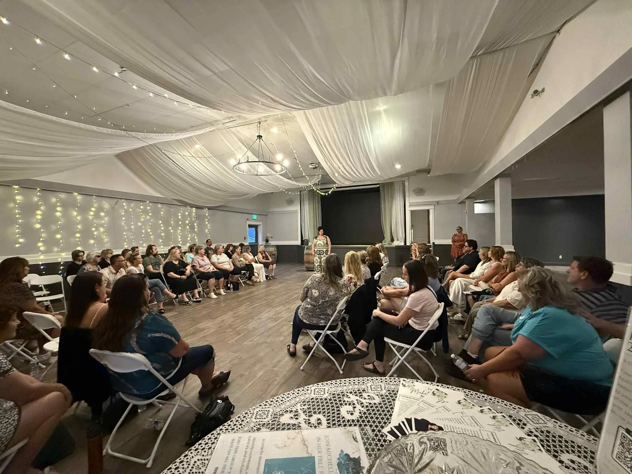 A group of people seated on white chairs in a large decorated indoor space, attending a presentation or event. The room has a draped ceiling with string lights and wall lights.