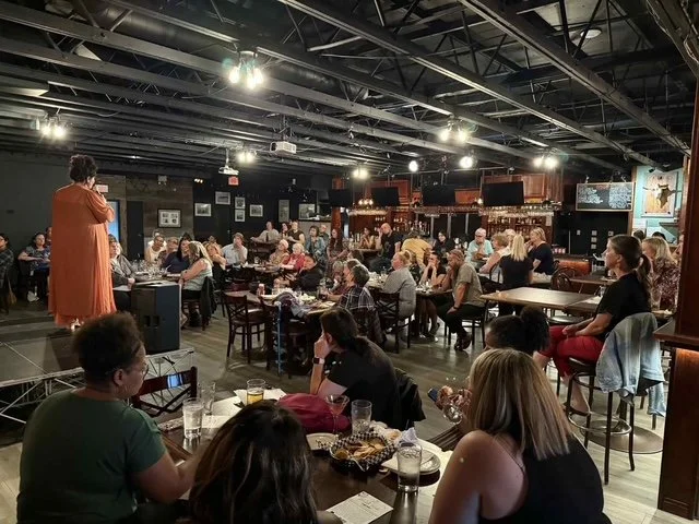 A woman in an orange dress performs on stage at a bustling bar or restaurant with many patrons seated at tables, watching her.