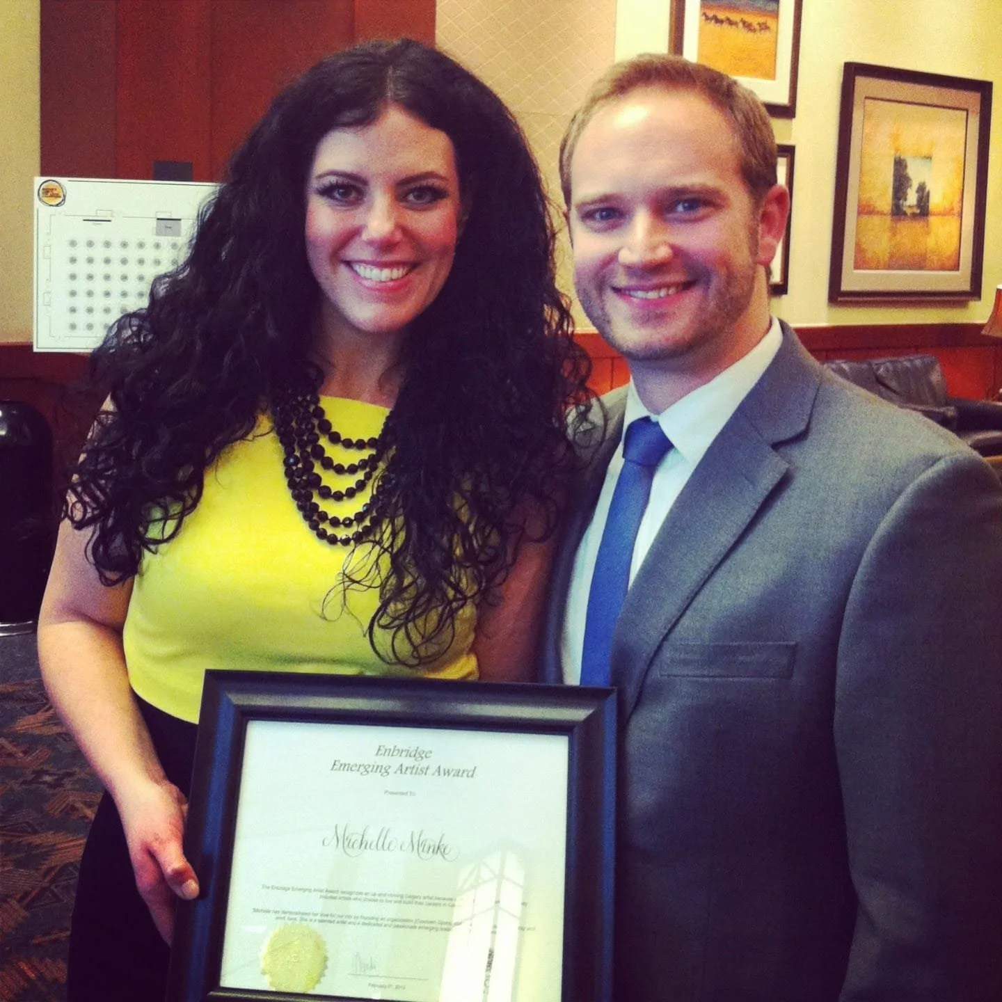 Woman in yellow dress holding an award certificate, standing next to a man in a gray suit. They are smiling and posing indoors with framed artwork on the wall behind them.