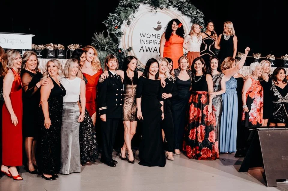 Group of women in formal attire at an awards event, standing on stage with a decorated backdrop and a sign that reads 'Women Inspiring Awards'.