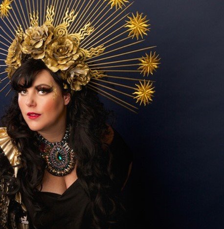 A woman wearing an elaborate gold floral headpiece with radiating gold spikes and star-like decorations, along with a statement necklace and black outfit, against a dark background.