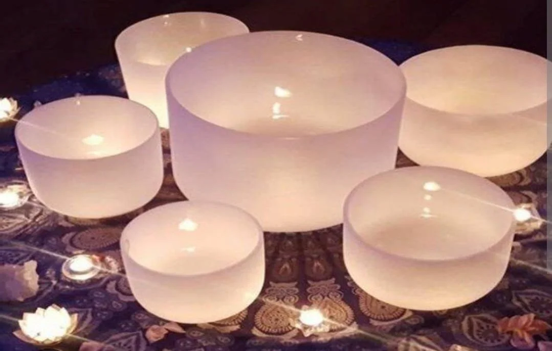 Seven white singing bowls of varying sizes arranged on a decorative cloth, illuminated by small candles around them.