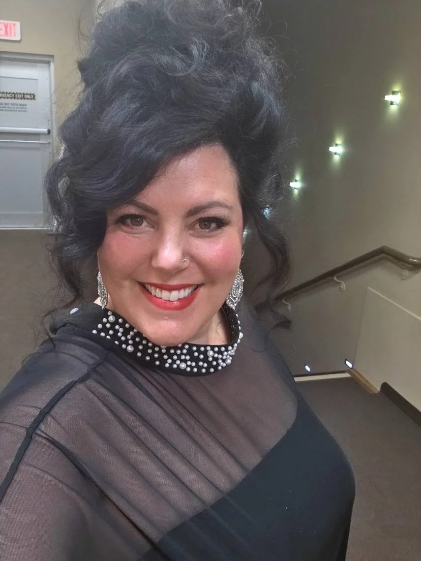 A woman with dark, curly hair styled in an updo, smiling in an indoor setting. She is wearing large, sparkly earrings and a black, sheer top with a beaded collar.