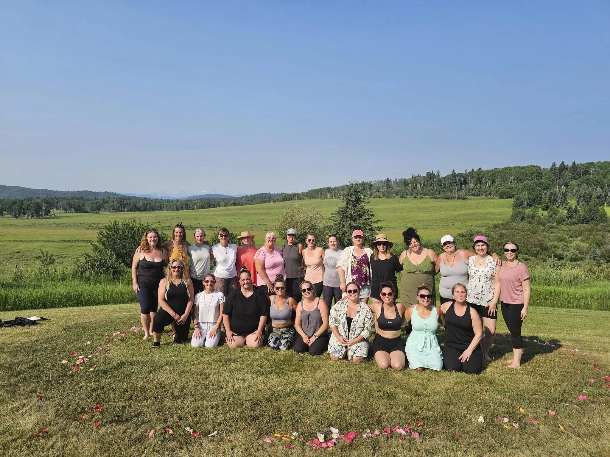 Group of women outdoors in a grassy field with green hills and trees in the background. They are smiling and posing for a photo, some wearing hats and sunglasses.