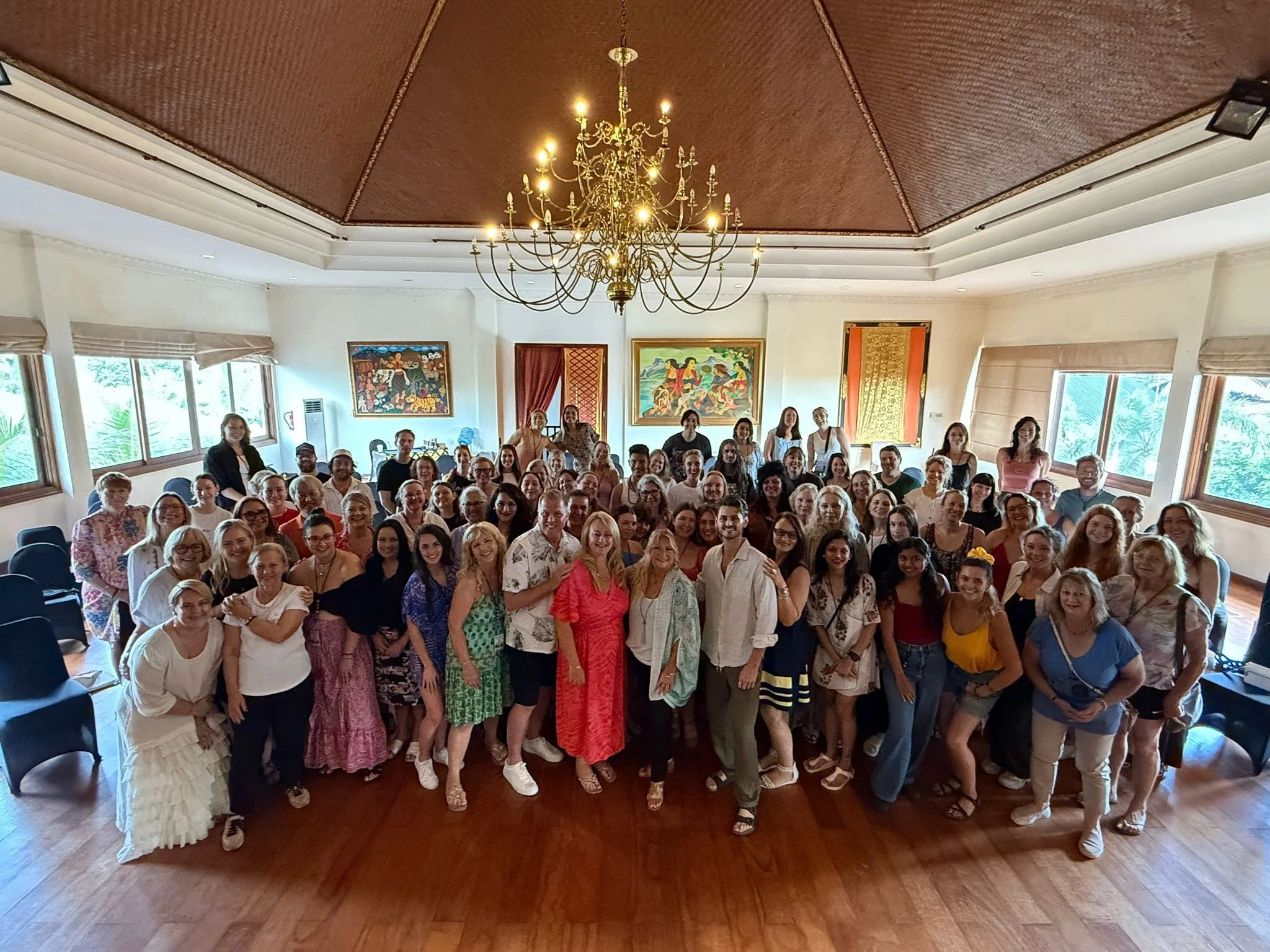 A large group of people gathered together in a spacious, well-lit room with wooden floors and large windows. The group includes men and women of various ages, smiling for the camera. The room features a chandelier hanging from the ceiling and vibrant
