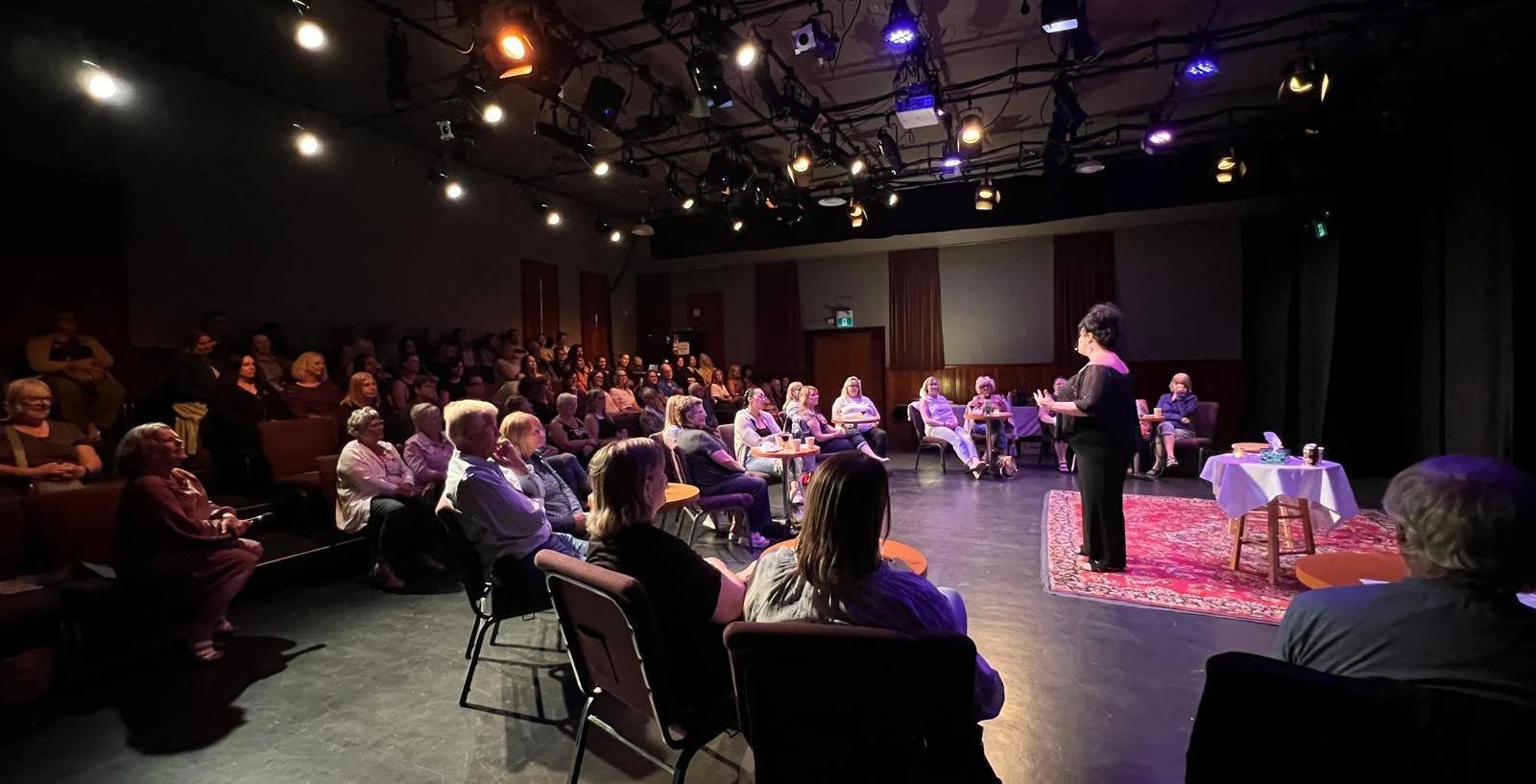 A woman speaking on stage to an audience seated in a theater or auditorium. The stage has a table with items and a red rug, with lighting overhead.