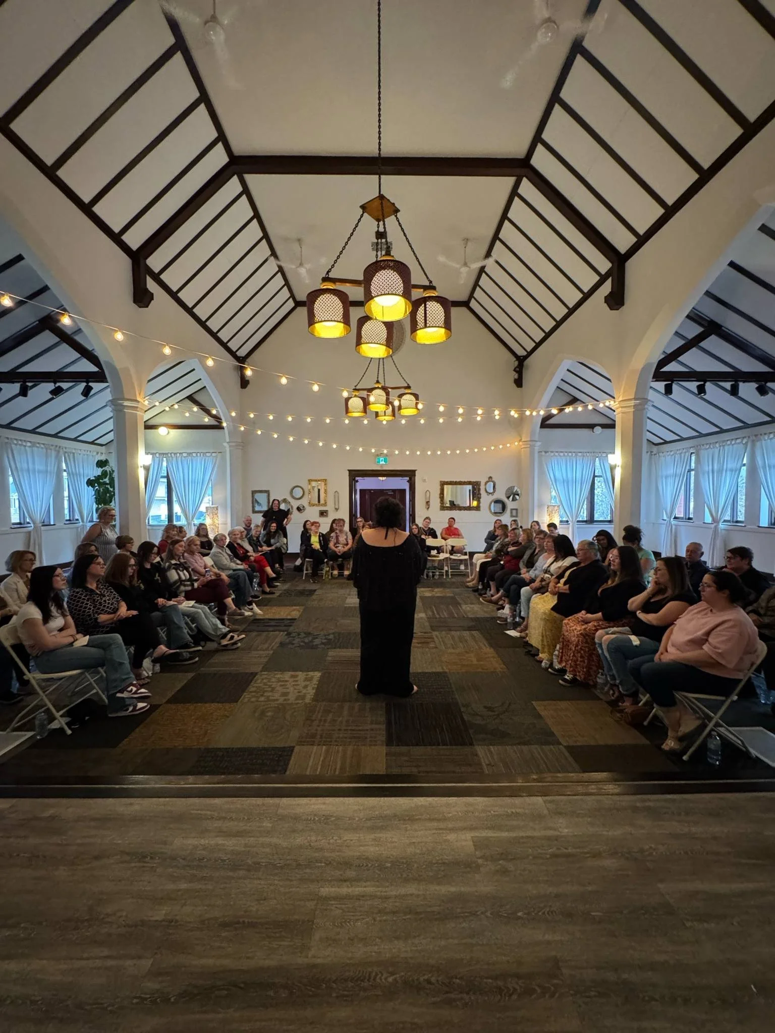 A large gathering in a decorated banquet hall with high, vaulted ceilings, string lights, and chandeliers, with people seated in a circle and a woman standing in the center.