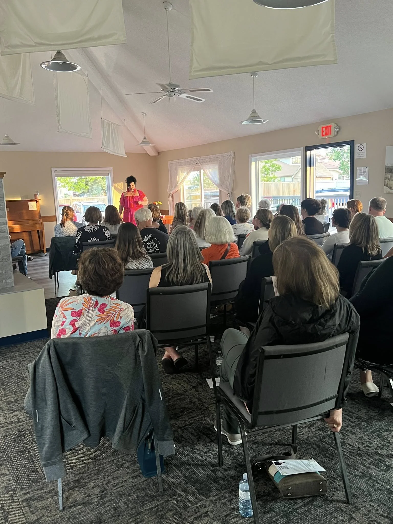 Woman in a red dress speaking to an audience seated in a room with large windows and white curtains.