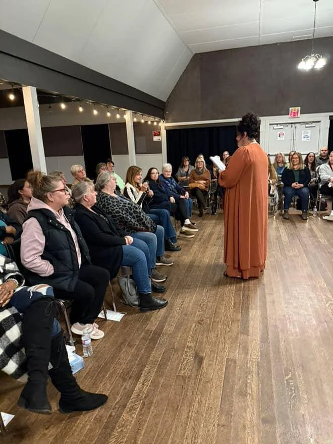 A woman in a long brown dress stands reading from a paper in front of a seated audience in a large room with wood flooring and dark walls, some audience members are clapping.