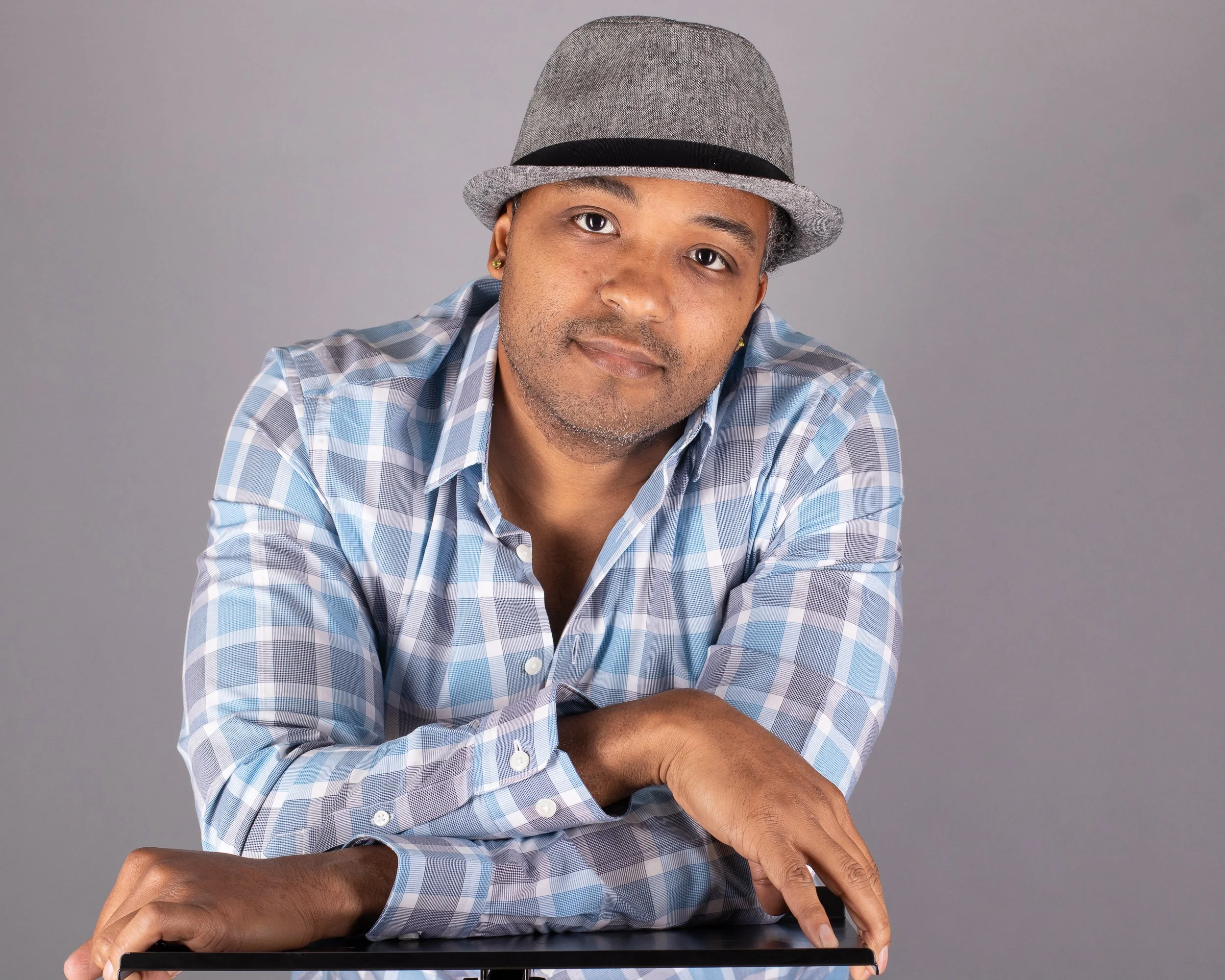 A man wearing a gray fedora hat and a blue plaid shirt, leaning on a surface, against a gray background.